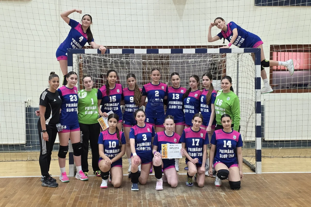 Fotografie de grup a echipei de Junioare 3: jucătoarele în echipament albastru cu roz, două cocoțate pe poartă, una ține diploma.