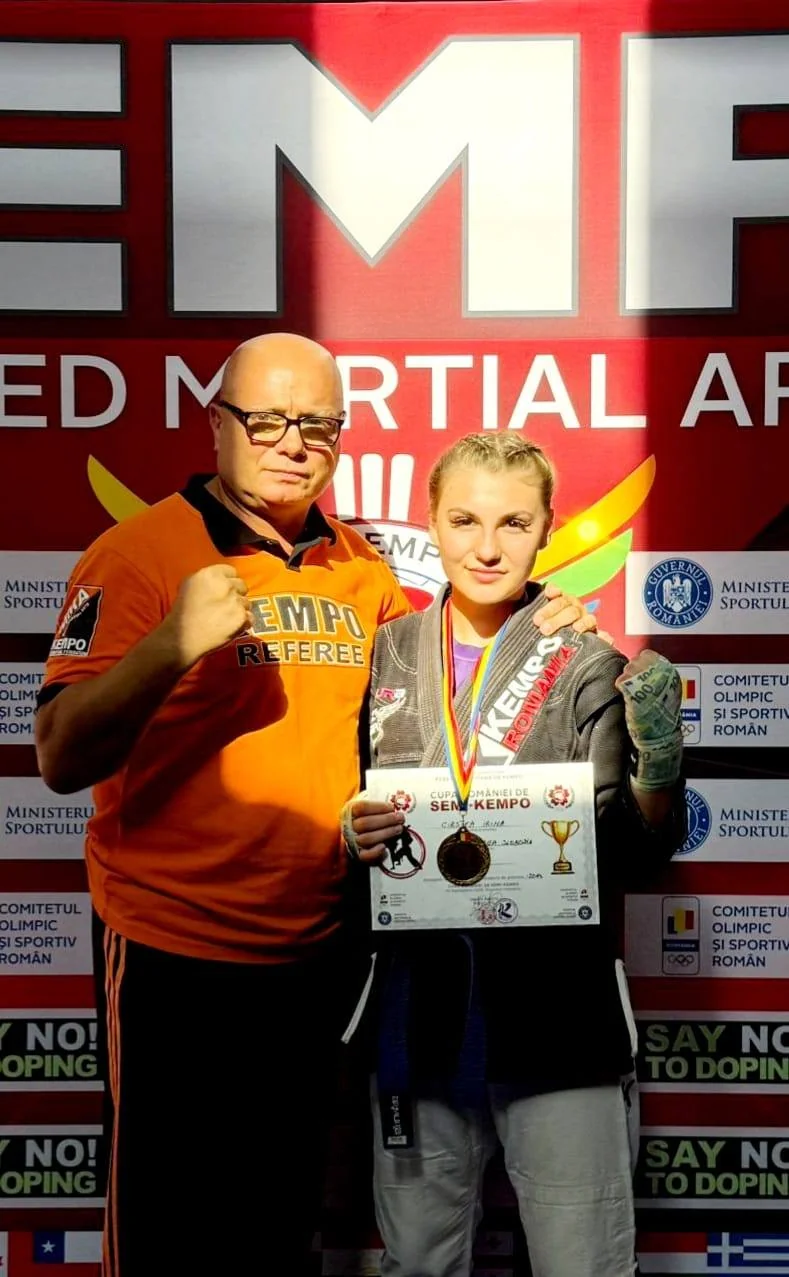 Irina Cîrstea arată medalia și diploma Cupei României la Semi-Kempo, lângă un arbitru, în fața panoului competiției.