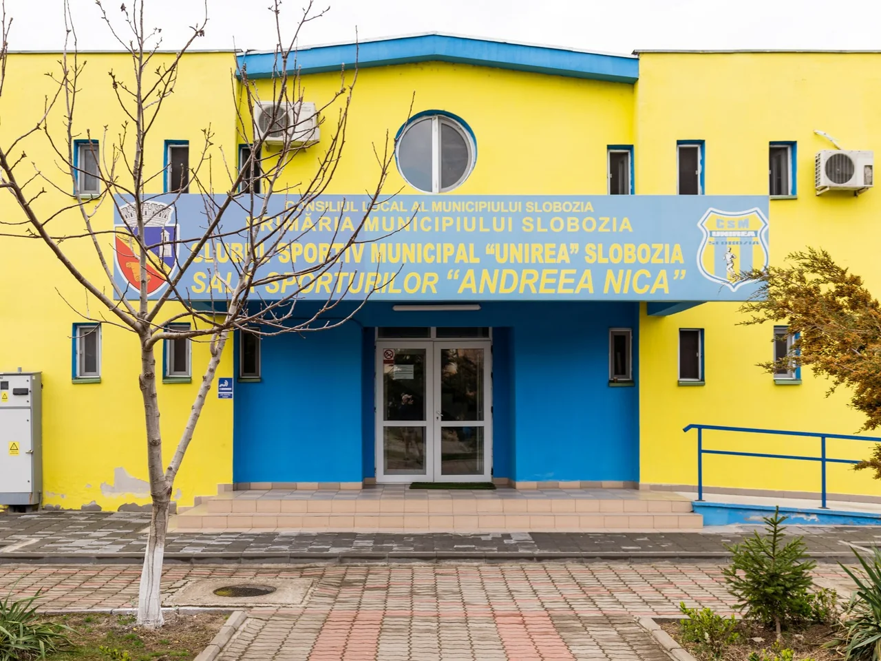 Intrarea Sălii Sporturilor „Andreea Nica”, o clădire galbenă cu albastru, cu numele sălii pe firmă.