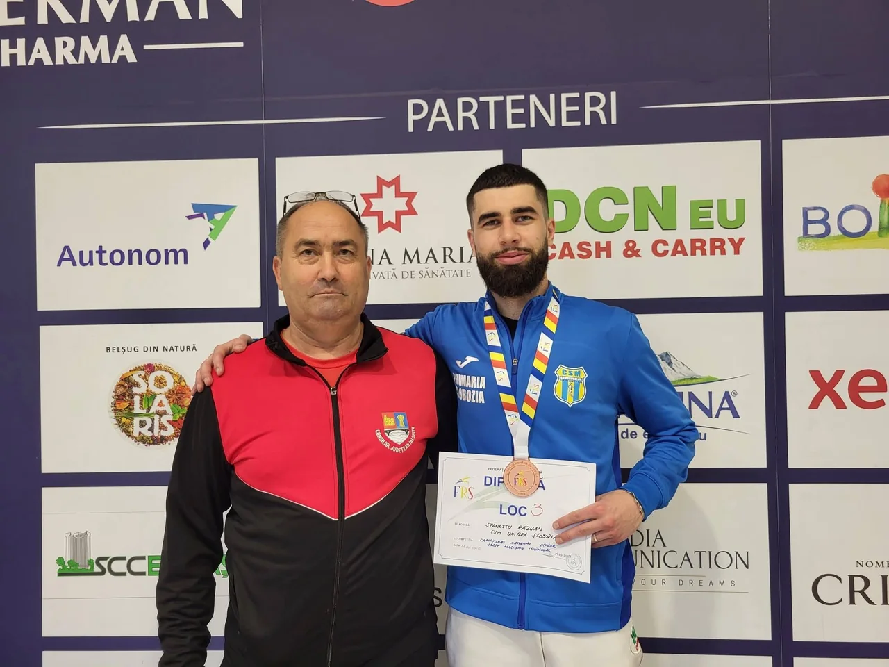Un sabrer al clubului, cu medalia de bronz și diploma, alături de antrenor, în fața panoului cu parteneri.