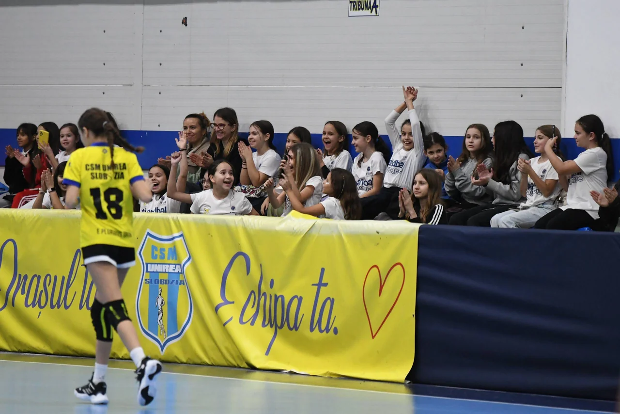 Tribuna plină de fete care aplaudă și strigă, deasupra bannerului „Orașul tău. Echipa ta.”; în față, o jucătoare în galben.