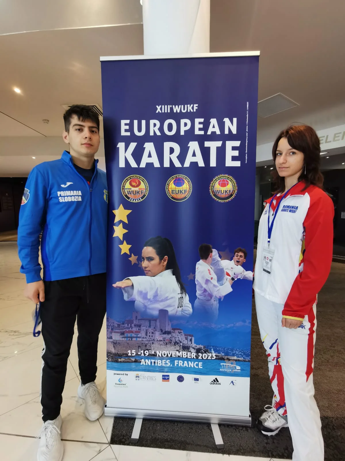 Un sportiv în geaca clubului și o sportivă în echipamentul României, lângă afișul Campionatului European de Karate WUKF de la Antibes.