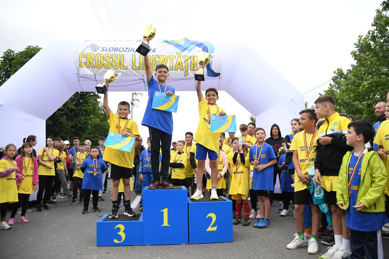 Trei băieți ridică cupele pe podiumul Crosului Libertății, înconjurați de alți participanți cu medalii.