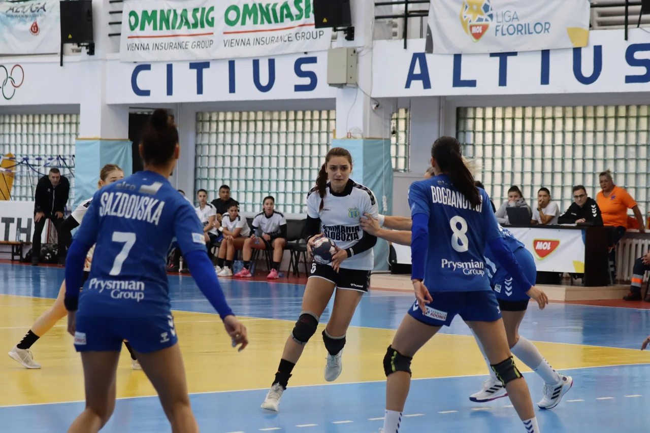 O jucătoare în alb, cu mingea în mâini, în fața apărătoarelor CSM Slatina, în albastru, în sala de la Slatina.