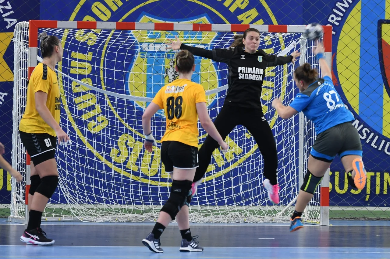 Meci de handbal feminin în Divizia A: o jucătoare aruncă la poartă din săritură, iar portarul în negru sare să apere.