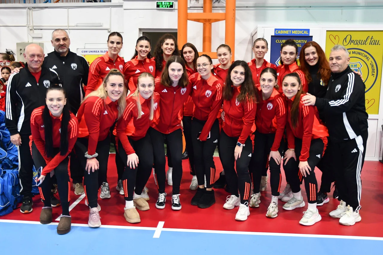 Echipa de handbal senioare, în treninguri roșii, pozează în sală alături de antrenori.