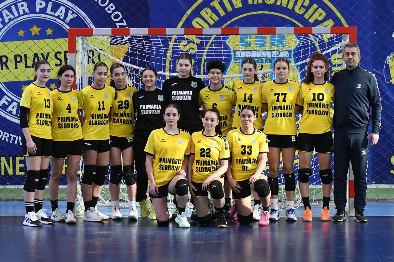 Echipa de junioare 2 de handbal, în galben, cu portarii în negru și antrenorul, în poză de grup în fața porții.