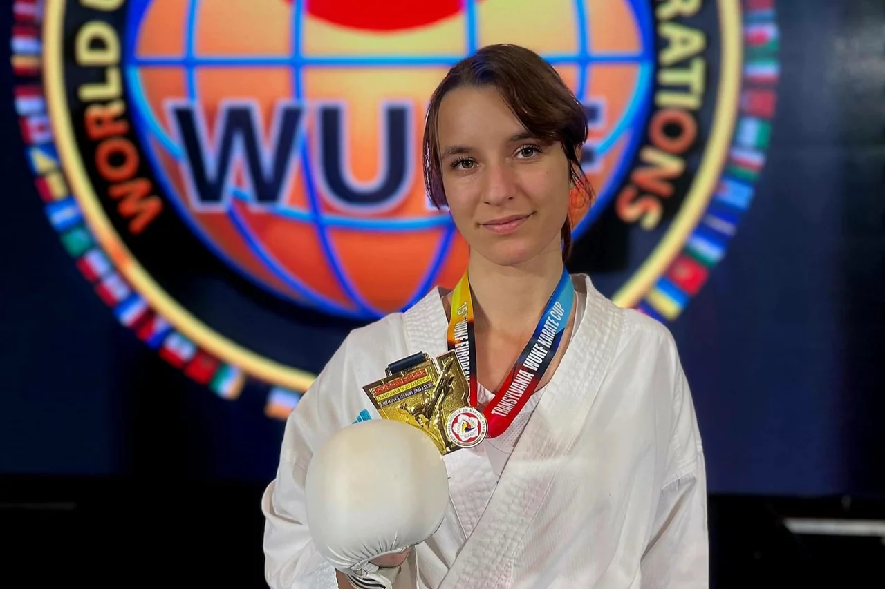 Ana Maria Matache, în kimono alb, cu medalia de aur și un trofeu, în fața siglei WUKF.