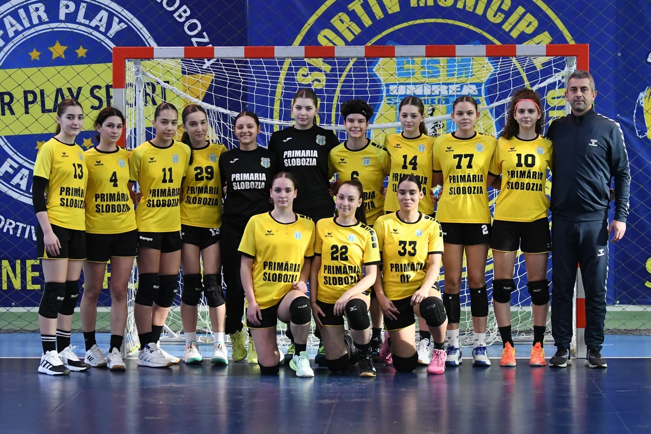 Echipa de junioare 2 de handbal, în galben, cu portarii în negru și antrenorul, în poză de grup în fața porții.