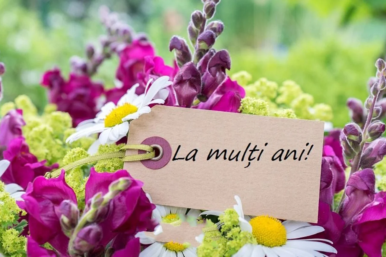 Buchet de flori mov, galbene și albe, cu o etichetă pe care scrie „La mulți ani!”.