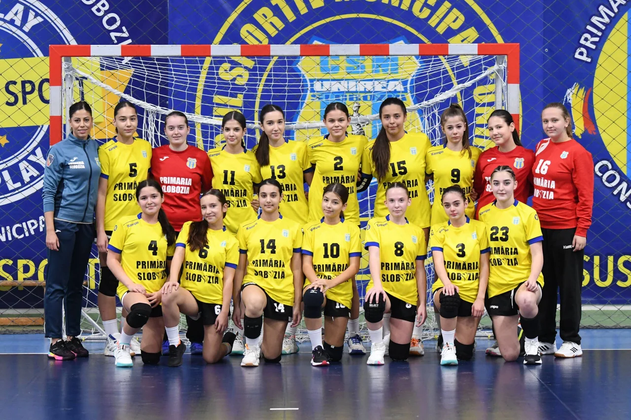 Echipa de junioare 3 de handbal, în echipament galben și cu portarii în roșu, în poză de grup cu antrenoarea în fața porții.