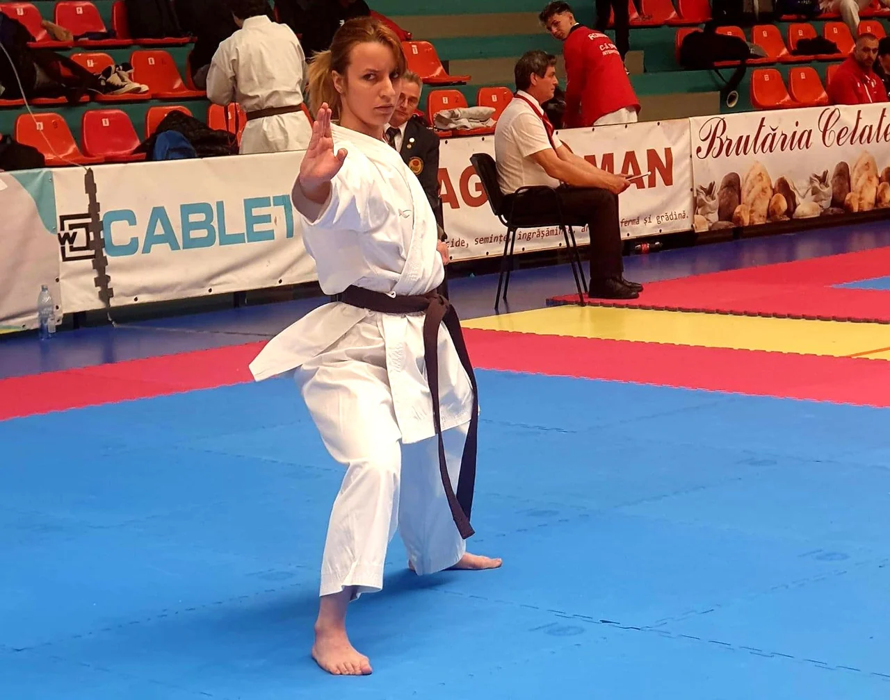 O karateka în kimono alb execută o kata pe tatami, cu arbitri și spectatori în fundal.