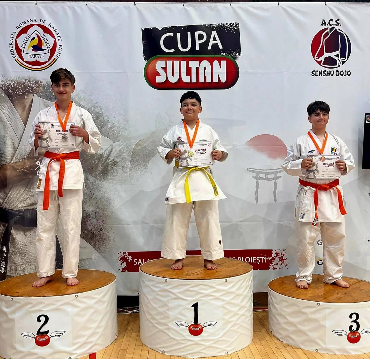 Trei băieți în kimonouri, cu medaliile la gât și diplomele, pe podiumul Cupei Sultan la karate.