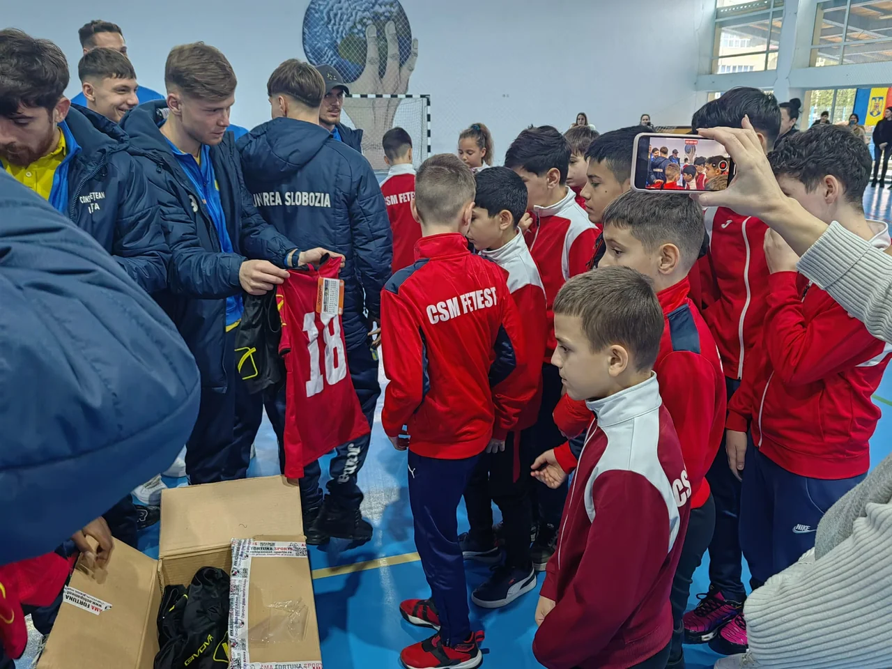 Fotbaliștii Unirii Slobozia împart copiilor de la CSM Fetești echipamente din cutii, iar un adult filmează cu telefonul.