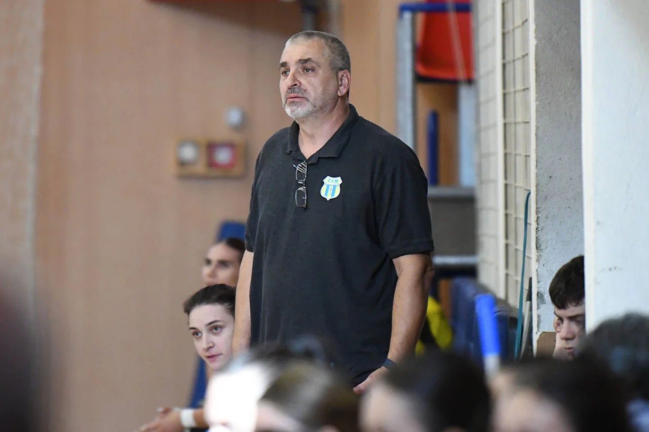 Antrenorul echipei de handbal, în tricou polo negru cu sigla clubului, urmărește jocul lângă banca de rezerve.