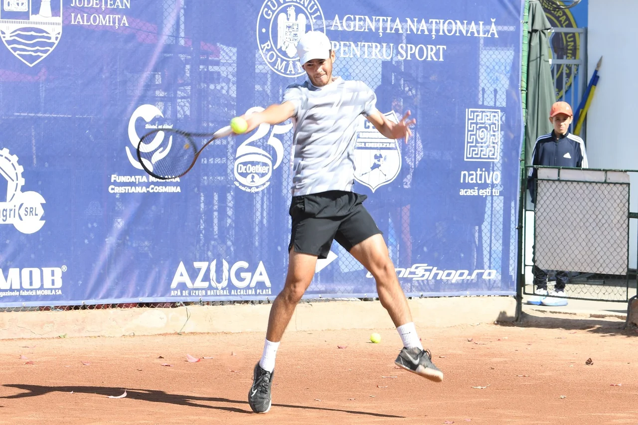 Ștefan Andreescu lovește mingea din forehand pe terenul de zgură, în fața unui panou cu sponsori.