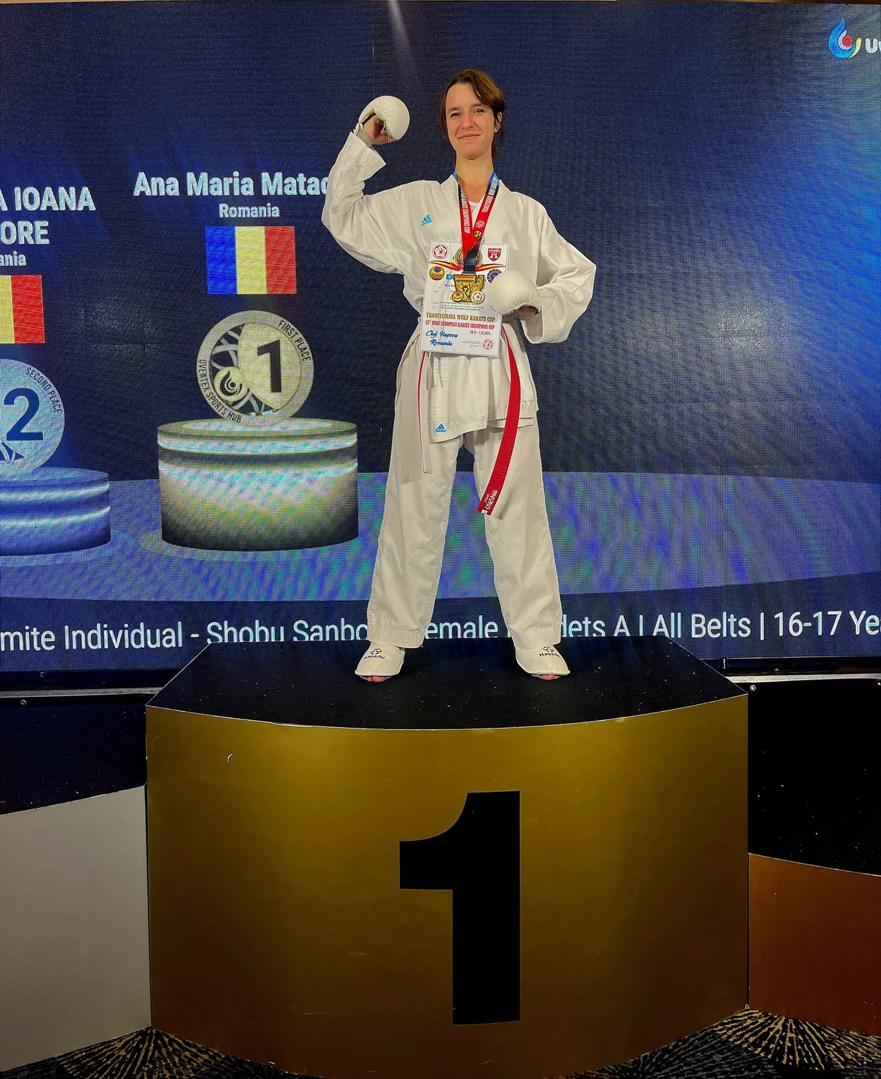 Ana Maria Matache pe prima treaptă a podiumului, cu ecranul „Kumite Individual, Female Cadets, 16-17 Years” în spate.
