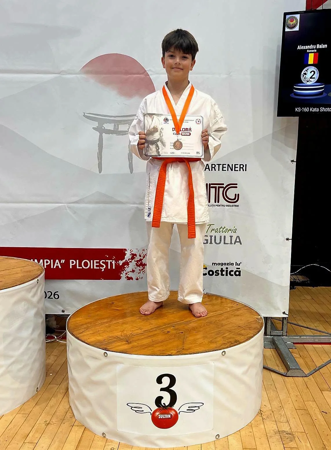 Un băiat în kimono alb cu centură portocalie, cu medalia și diploma, pe treapta a treia a podiumului.