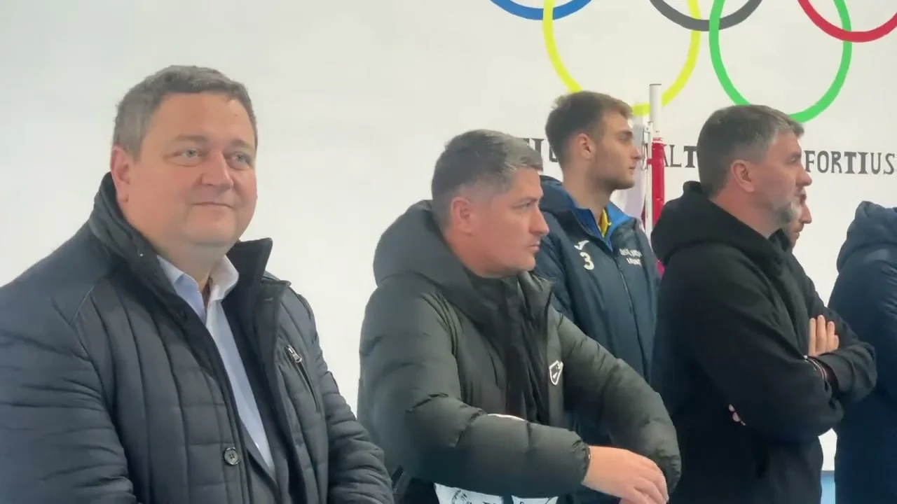 Patru bărbați urmăresc acțiunea de donare a echipamentelor, în sala de sport cu cercurile olimpice pe perete.