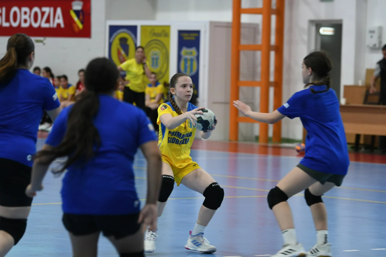 Jucătoare a echipei de junioare 3, în galben, cu mingea în mâini în fața apărătoarelor de la CSS Ilfov, în albastru.