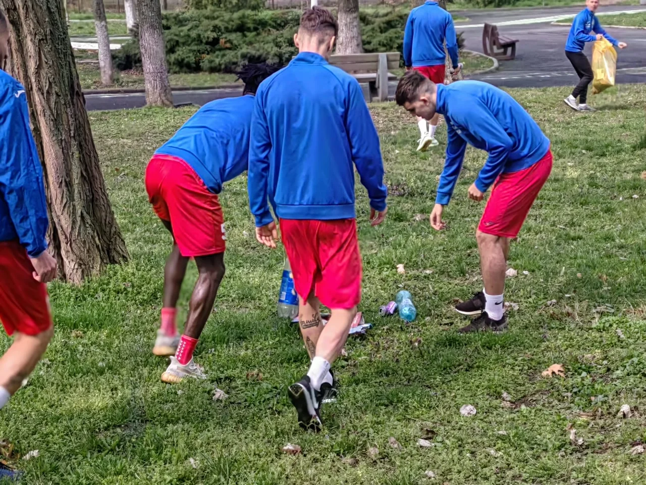 Fotbaliști în bluze albastre și șorturi roșii strâng de pe iarbă sticle și gunoaie, în parcul de lângă stadion.