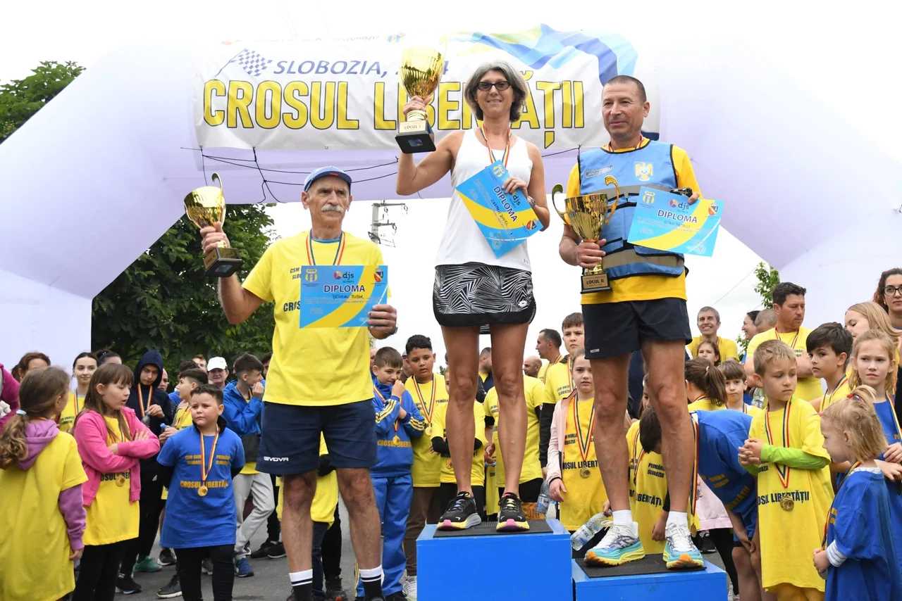 Premierea alergătorilor adulți la Crosul Libertății: trei câștigători cu cupe și diplome pe podium, cu mulți copii în jur.
