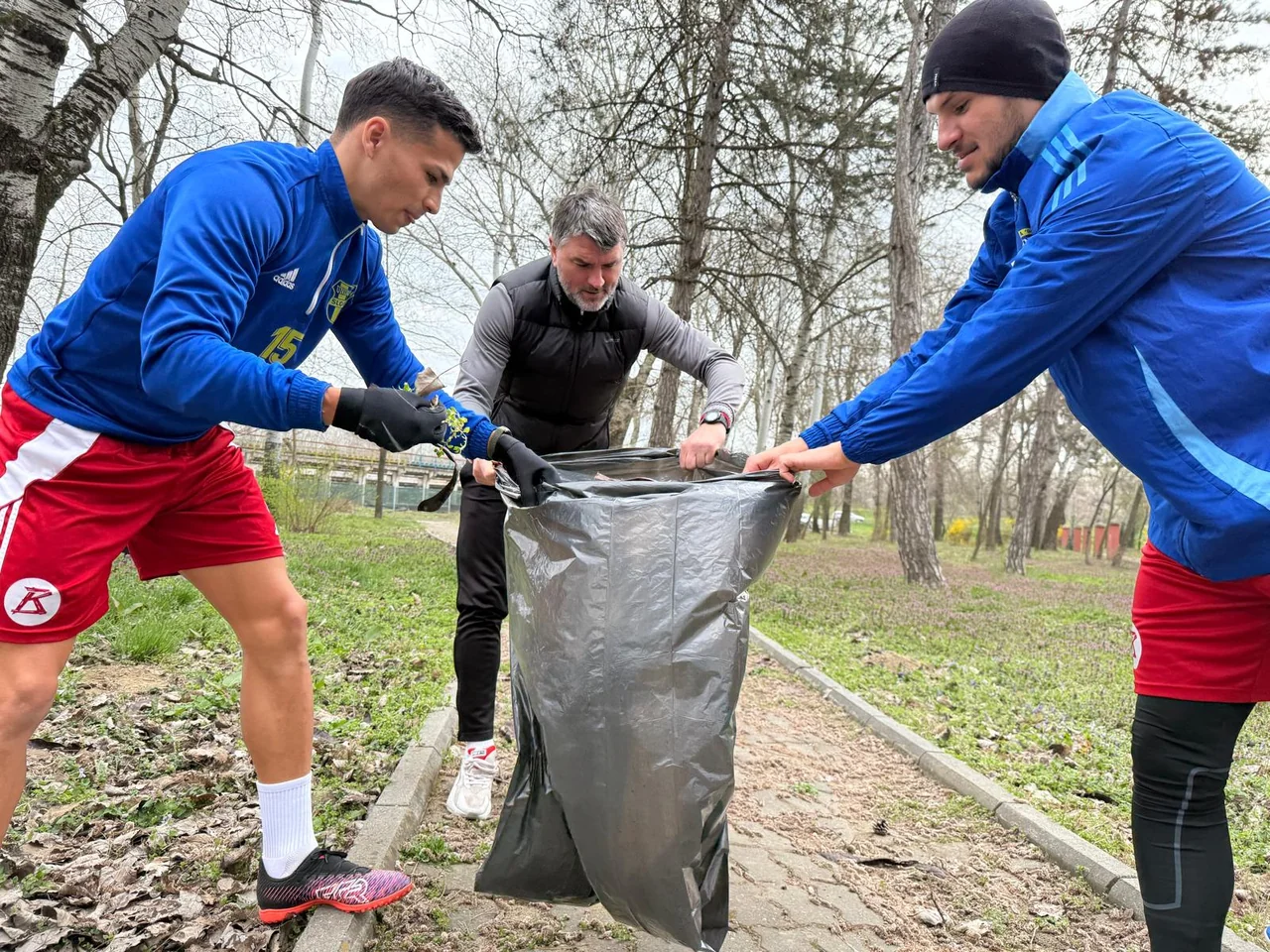 Doi fotbaliști și un membru al staff-ului țin un sac de gunoi la acțiunea de ecologizare din parc.