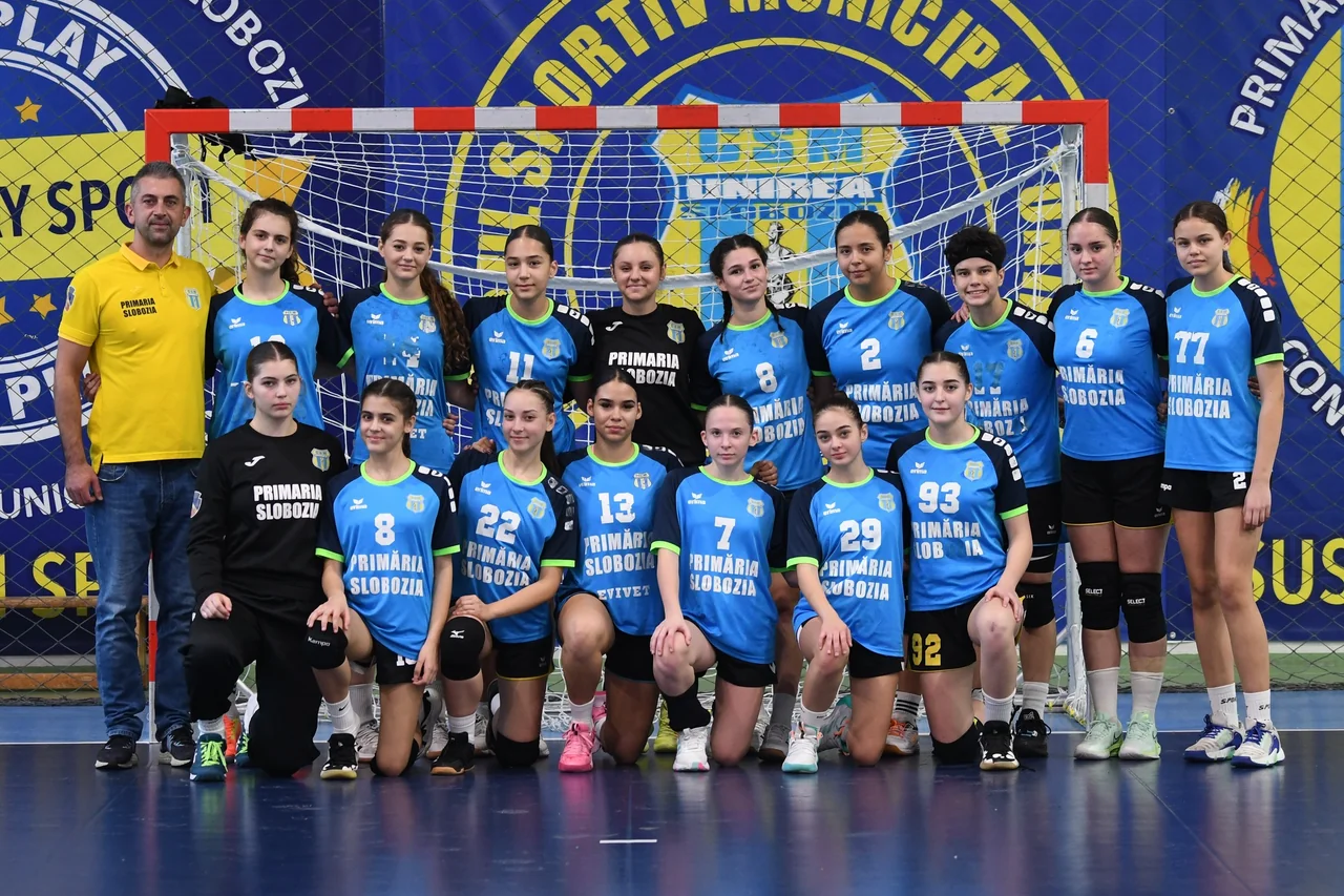 Echipa de junioare 2 de handbal, în echipament albastru, în poză de grup cu antrenorul în fața porții.