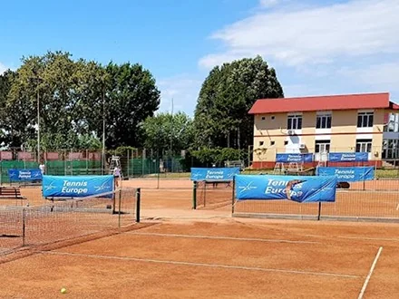 Terenurile de tenis cu zgură, cu fileele și bannerele Tennis Europe, iar în fund clădirea bazei.