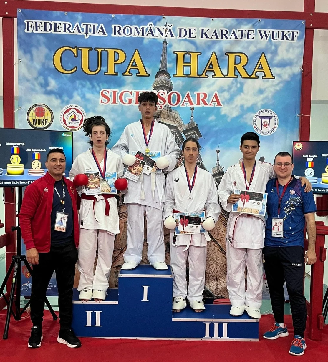 Karateka medaliați pe podium, cu diplome, și doi antrenori, sub bannerul „Cupa Hara, Sighișoara”.