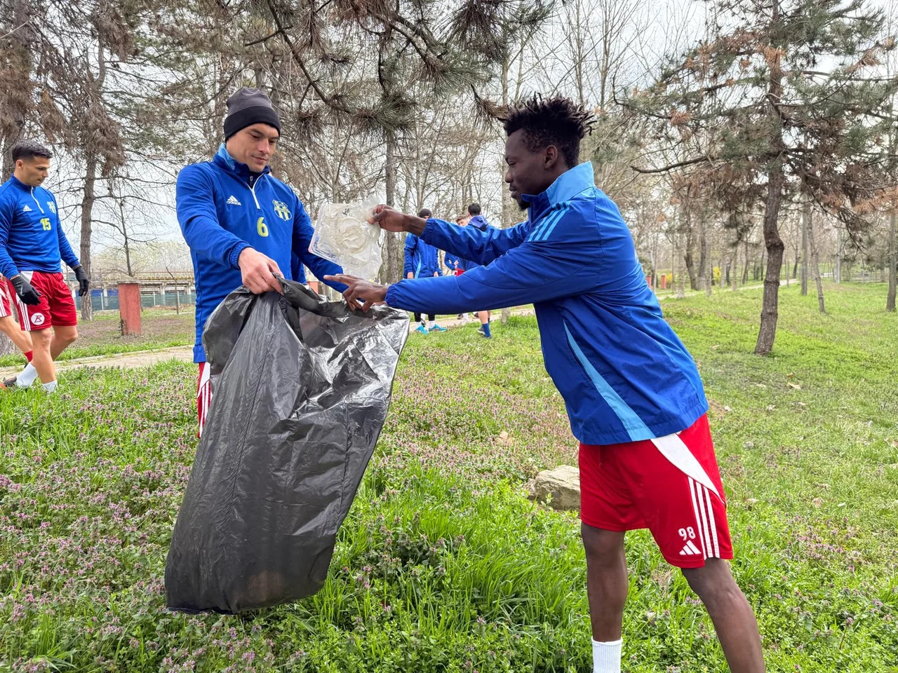 Doi fotbaliști Unirea Slobozia pun gunoaie într-un sac negru, pe iarba din parc.